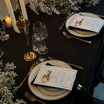 Elegant wedding table with a black tablecloth, golden cutlery, crystal glasses, and personalized nameplate 'Bride' on the plate, surrounded by flowers and a menu card.